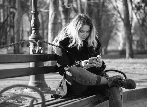 Black and white photo of a girl in a coat in a park sits in an interesting pose on a bench with a phone in her hands. Social Media Concept - Φωτογραφία, εικόνα