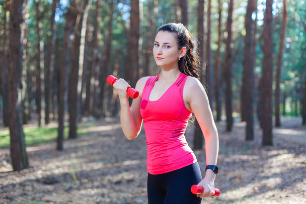 Sportive attractive girl working out with dumbbells, training in the forest. Fitness, sport, lifestyle concept - Photo, Image
