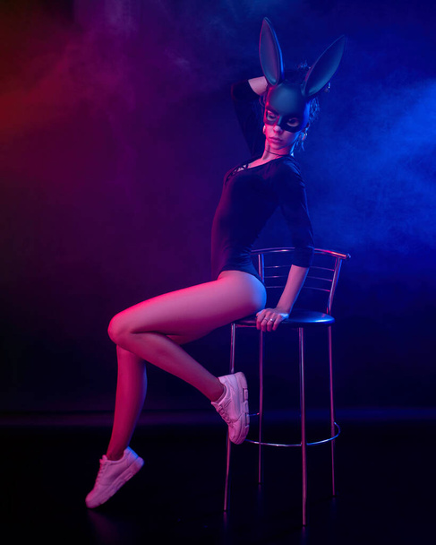 the beautiful girl in a bodysuit poses in a photo Studio on a dark background in neon light. On the face of a rabbit mask - Foto, Bild