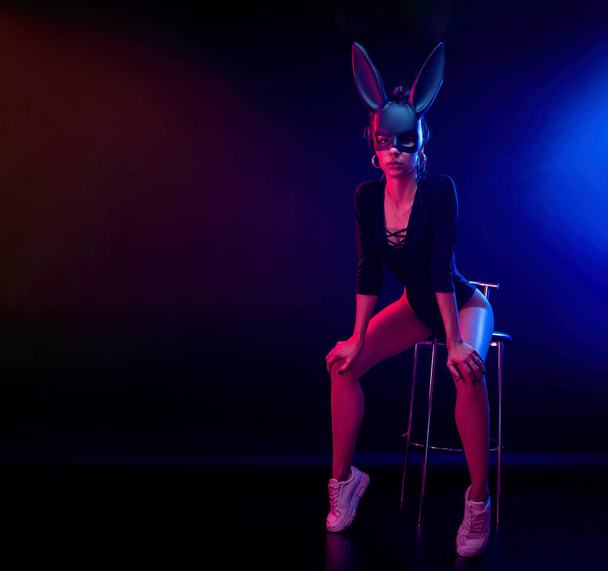 Vücut giysisi giymiş güzel bir kız, bir fotoğraf stüdyosunda, neon ışıkta, karanlık bir arka planda poz veriyor. Tavşan maskesinin yüzünde - Fotoğraf, Görsel