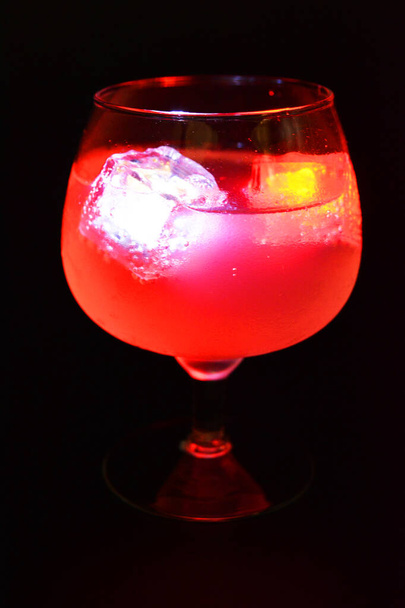 White and red bright glowing ice cubes that float in a delicious frosted white cold drink are located in a cognac glass on a black frosted background. - Foto, Imagem