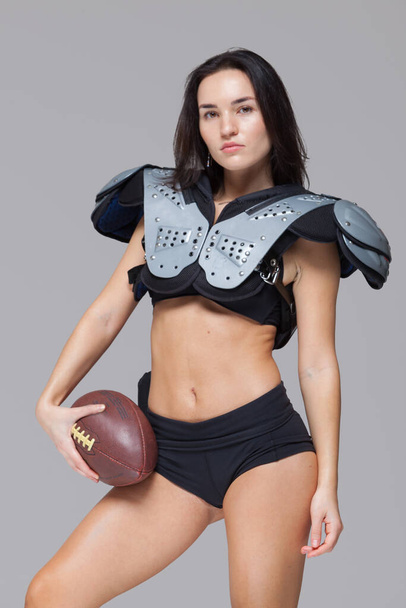 Young sporty brunette wearing sexy uniform of American football player posing with ball looking at camera isolated on grey background - Foto, immagini