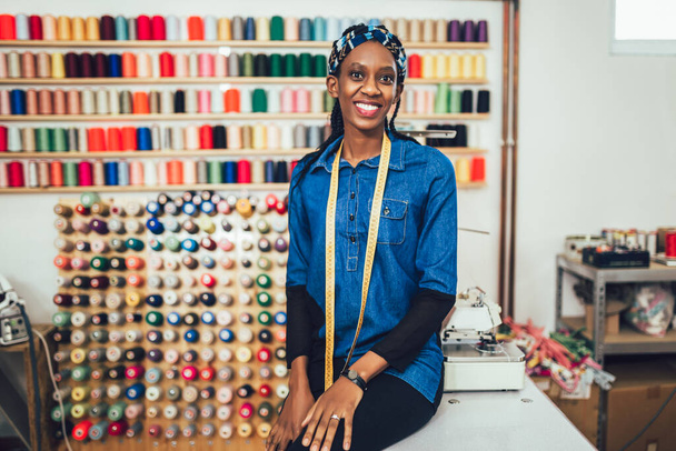 Portret van een vrolijke Afrikaanse kleermaakster in de studio. Achtergrond van kleurrijke naaigaren. - Foto, afbeelding