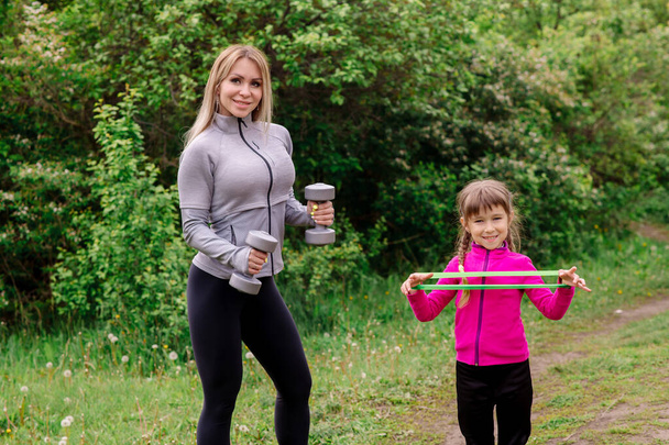 Mom and daughter 5 years old in sportswear are training in the park with dumbbells and expanders - Фото, изображение