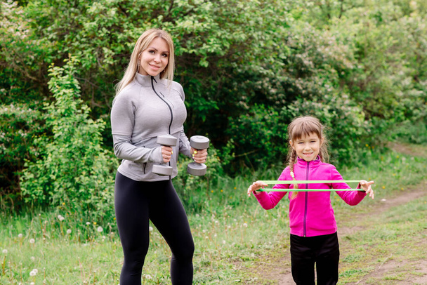 Mom and daughter 5 years old in sportswear are training in the park with dumbbells and expanders - Foto, afbeelding
