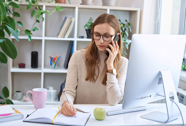 Fiatal elfoglalt üzletasszony, vonzó női alkalmazott szemüvegben ül az asztalnál, telefonon beszél, és jegyzetel, miközben távolról dolgozik otthon - Fotó, kép
