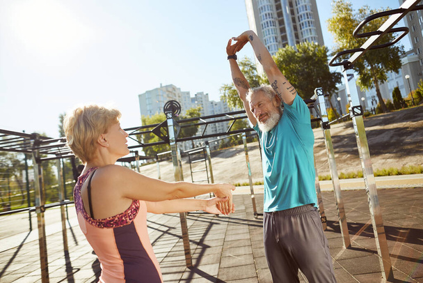Staying healthy. Active mature family couple in sportswear warming up together at outdoor gym in the morning, aged couple doing stretching exercises outdoors - Photo, Image