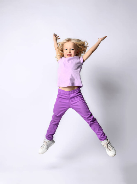 Little blonde kid in purple blouse and pants, white sneakers. Smiling, jumping up with raised hands, posing isolated on white - Photo, Image