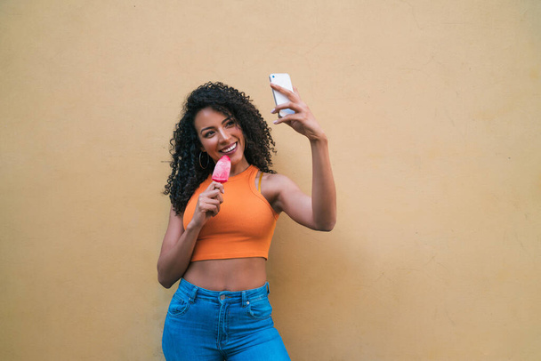 Portrait of afro woman taking selfies with her mophile phone while eating ice-cream. Technology and lifestyle concept. - Foto, afbeelding