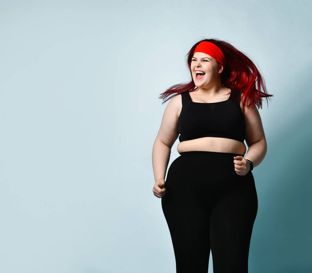 Active young plus-size woman with flowing red hair in black sport top and leggings shouting with laughter and delight on blue background. Copy space - Foto, Bild