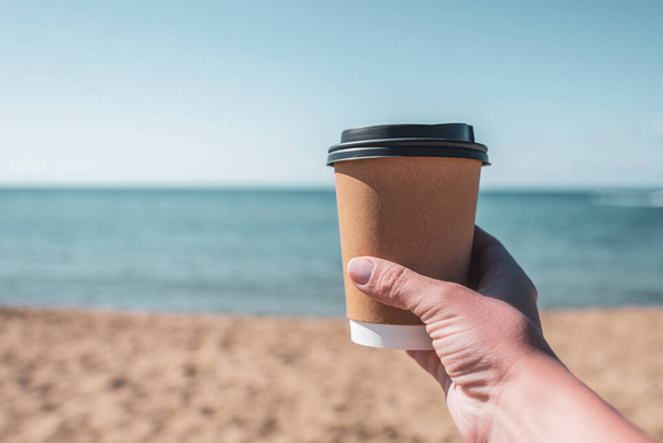 Paper mug, cup of coffee in woman hand, sand of sunny beach opposite the sea wave. summer. take away. breakfast - Foto, Imagen