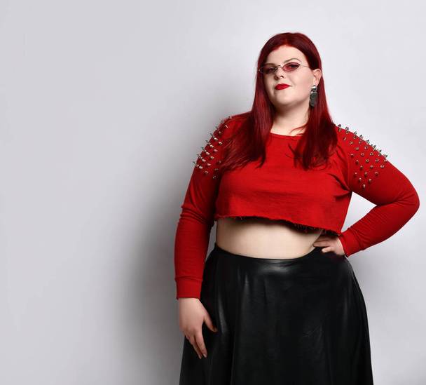 Obese redhead lady in red spiked top, black skirt, sunglasses. She has put hand on waist, posing isolated on white background - Foto, Bild
