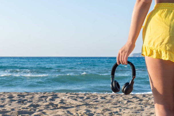 Relaxed woman walking on the beach with headphones meditating listening to music at sunset or sunrise. Girl in the yellow summer suit, sand, sea view. - Photo, Image