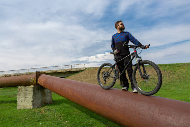 Ciclista barbudo en una bicicleta de montaña en una tubería oxidada, hierba verde, cielo azul
 - Foto, imagen