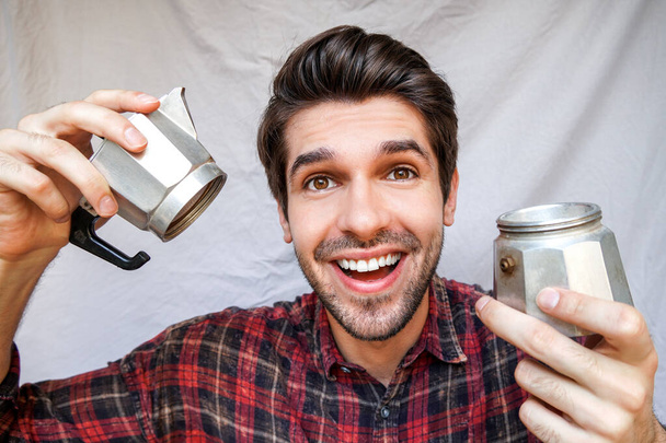 Portret van een jonge vrolijke man met een grote glimlach die een open glimmende moka pot vasthoudt met een koffie in een shirt - Foto, afbeelding