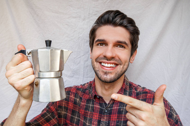 Portret młodego, wesołego mężczyzny z uśmiechniętą twarzą wskazującą na błyszczący moka pot z kawą w koszuli - Zdjęcie, obraz