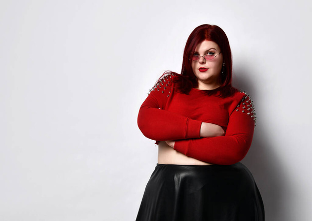 Overweight red-haired woman in a red studded top, black leather skirt and earrings. She stands with her arms folded in a twisted pose - Fotó, kép