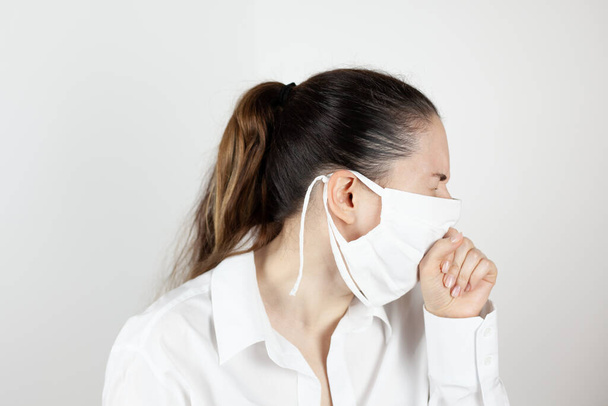 A young girl in a mask coughs and covers her face with her hand out of habit. Demonstration of habits. Life during the pandemic. The first signs of the disease. - 写真・画像