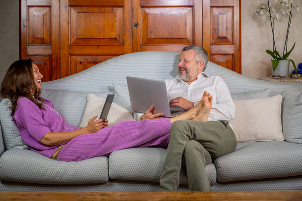 La feliz pareja se ríe y bromea mientras trabaja en casa en el sofá con el ordenador portátil y la tableta - La esposa está trabajando en el sofá y el marido está jugando con el ordenador
 - Foto, imagen