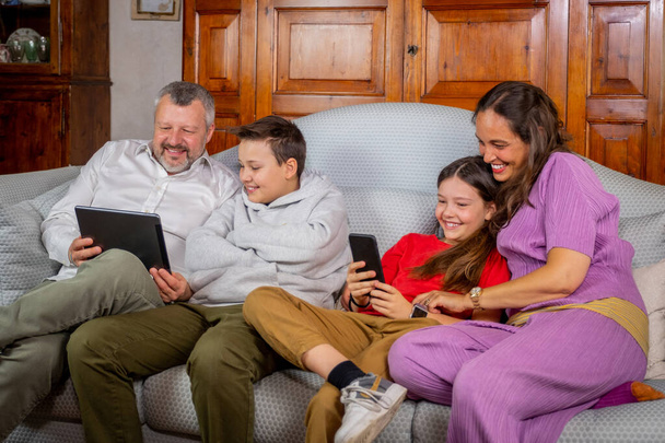 Familia feliz sentado en el sofá y el uso de la tableta digital y el teléfono móvil en casa - Padre e hijo riendo mientras ve tha tableta y la madre y la hija bromeando en el chat de vídeo con el teléfono inteligente
 - Foto, Imagen