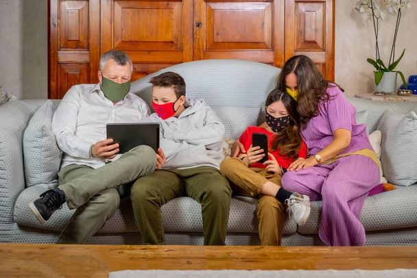 Mamma e papà guardano con i loro figli il tablet e lo smarphone sul divano di casa - Bella famiglia che indossa maschere facciali contro la pandemia mondiale coronavirus e stare a casa
 - Foto, immagini