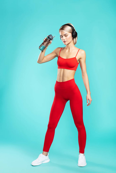 Egy kis szünet. Fiatal és sportos szőke nő fülhallgatóban és piros sportruházatban ivóvíz, miközben kék háttérrel szemben áll. Teljes hossz - Fotó, kép