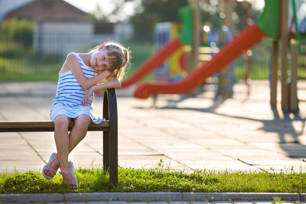 Schattig jong meisje in korte jurk zittend alleen buitenshuis op Speeltuin Bench op zonnige zomerdag. - Foto, afbeelding