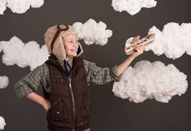 dívka si hraje s papírovým letadlem a sní o tom, že se stane pilotem, oblečená v retro bundě a helmě s brýlemi, mraky bavlněné vlny, šedé pozadí, tónovaná v hnědé - Fotografie, Obrázek