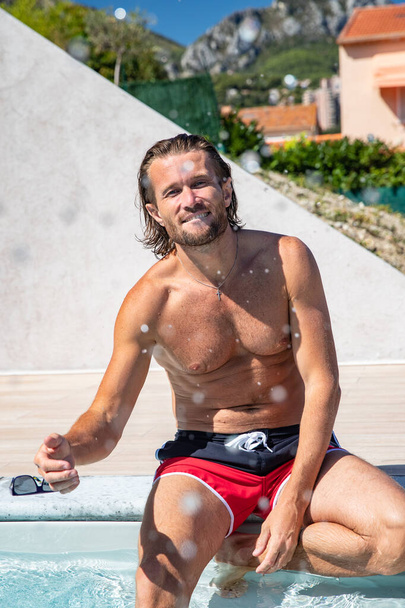 The handsome brutal man with a long hair and naked torso sits near the pool, splashes with water, he has fun, a sports suntanned body, he is red blue swimming shorts, sunny day - Фото, изображение