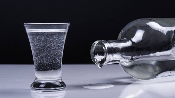 Un vaso de vodka y una botella reclinable con una gota sobre una mesa blanca y sobre un fondo negro
 - Foto, imagen