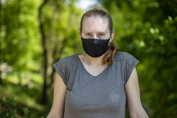 Portrait of a beautiful young woman in a black mask on a green background. Woman in a park during an epidemic, a coronavirus pandemic. Covid-19. - 写真・画像