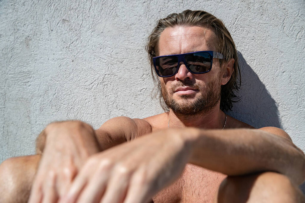 The handsome brutal man in sunglasses with a long hair and naked torso sits near the pool, is looking a camera, a sports suntanned body, sunglasses with a blue frame, he is red blue swimming shorts - Фото, изображение
