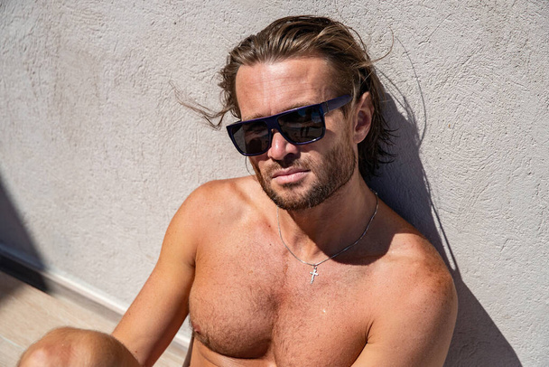 The handsome brutal man in sunglasses with a long hair and naked torso sits near the pool, is looking a camera, a sports suntanned body, sunglasses with a blue frame, he is red blue swimming shorts - Zdjęcie, obraz