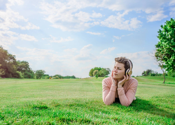 Woman in pink coat, black top and jeans listens to music / audiobook and rest on meadow / green grass on sunny spring warm day in park - Photo, Image
