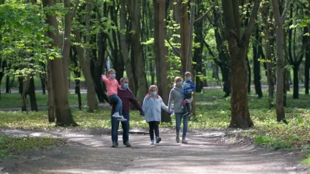 Sağlık maskeli bir aile ilkbaharda parkta yürür. Anne ve baba, çocukları kollarında taşıyorlar. Covid-19 koronavirüs enfeksiyonlarından korunma kavramı. - Video, Çekim