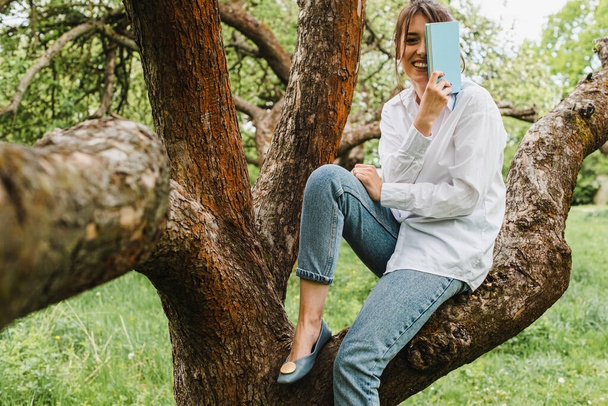 young girl in a white shirt and blue jeans sitting in nature on an apple tree reading a book. Self-isolation. Quarantine concept. Virus. Pandemic. Coronavirus - Photo, Image