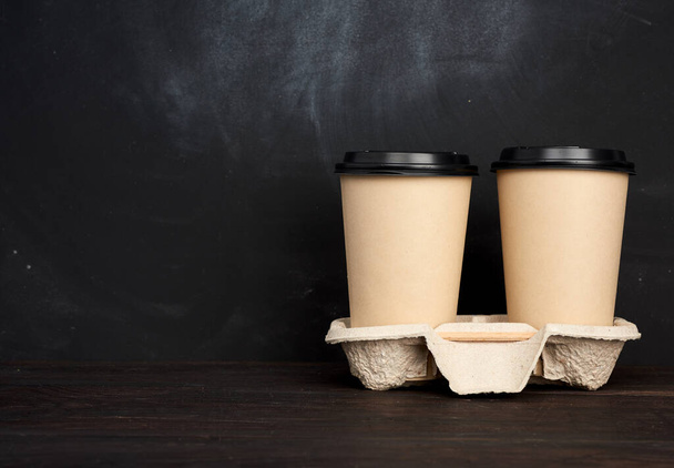 two brown paper disposable cups with a plastic lid stand in the tray on a wooden table, black background, place for an inscription, takeaway containers - Photo, Image