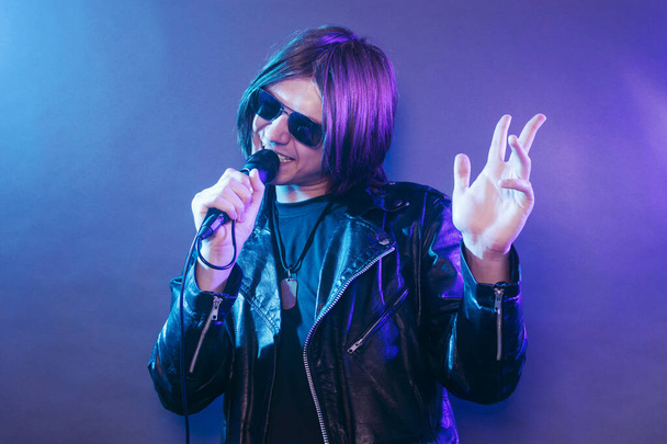 Mladá rocková hvězda v černé kůži zpívá v mikrofonu na modrém purpurovém pozadí. - Fotografie, Obrázek