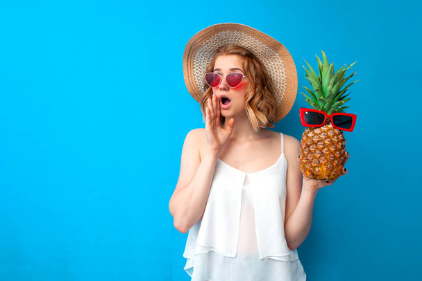 mladá dívka s ananasem v slunečním klobouku a brýlemi mluví na modrém pozadí, žena v letním oblečení křičí na místo pro text, letní koncept - Fotografie, Obrázek