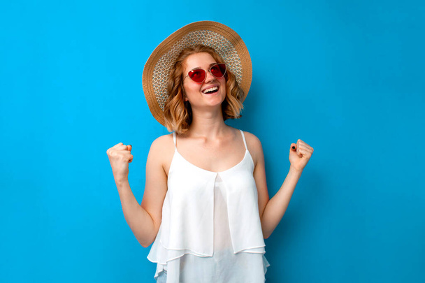 νεαρή κοπέλα σε ένα καπέλο ήλιο και γυαλιά χαίρεται τη νίκη σε ένα μπλε απομονωμένο φόντο, χαρούμενη γυναίκα δείχνει μια χειρονομία νίκης σε καλοκαιρινά ρούχα, έννοια του καλοκαιριού - Φωτογραφία, εικόνα