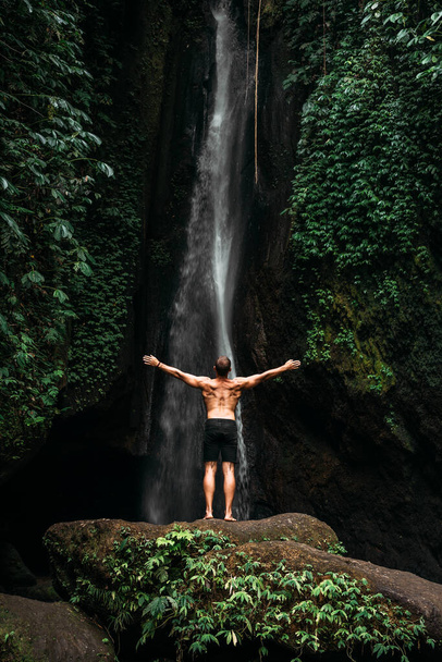 Back view of a man standing by waterfalls with arms outstretched. Man at waterfall raising his hands in feeling closer to nature. Man at the waterfall. Travel to Bali. Man on the background of nature. With copy space - 写真・画像