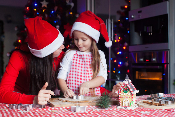 Adorable little girl with mother wearing santa hats baking Christmas gingerbread cookies together - 写真・画像