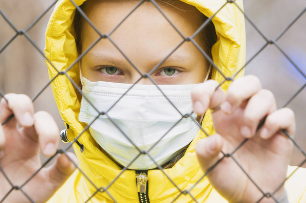 Tıbbi maskeli karantina altındaki kız çitin metal ağından bakıyor. - Fotoğraf, Görsel