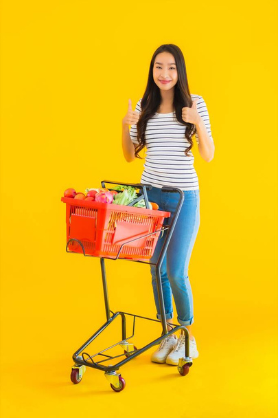 Portret mooie jonge aziatische vrouw winkelen supermarkt en kar op geel geïsoleerde achtergrond - Foto, afbeelding