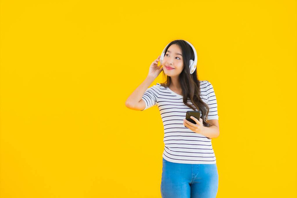 Retrato hermosa joven asiática mujer uso inteligente teléfono móvil con auriculares para escuchar música en amarillo aislado fondo
 - Foto, Imagen