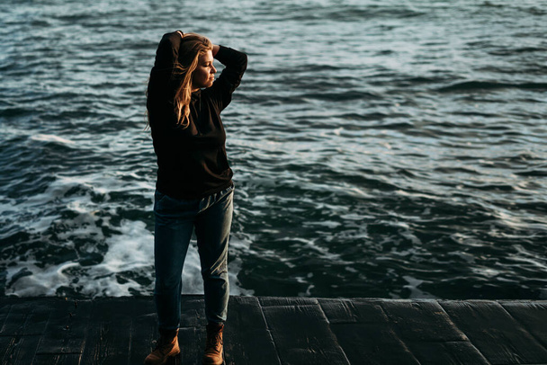  Retrato de crescimento completo de uma menina bonita jovem no cais junto ao mar e olha para o amanhecer em uma jaqueta preta e jeans, sapatos vermelhos. Mãos na cabeça. Paz, liberdade, realização da vida
 - Foto, Imagem