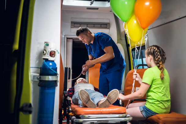 Un homme en uniforme médical met un masque à oxygène sur une fille allongée sur une civière, sa petite sœur regarde le travail du médecin
 - Photo, image