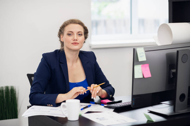 Бизнес-концепция. Портрет улыбающейся молодой привлекательной деловой женщины, сидящей за столом и смотрящей в камеру
 - Фото, изображение