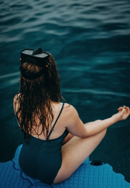 Bulanık kız suya yakın bir rıhtımda turkuaz bir mayoyla dalış maskesiyle oturur ve meditasyon yapar. Arkadan bak, yakın plan. Açık mavi deniz. İç huzuru. Yaşamın yansımaları. Rahatlama - Fotoğraf, Görsel