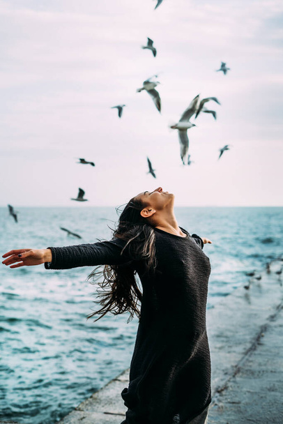 Close-up A young girl in black clothes is standing by the sea with a strong wind. seagulls fly. The power of thought, magic, self-awareness, gaining strength to overcome difficulties. Mental health - Photo, Image